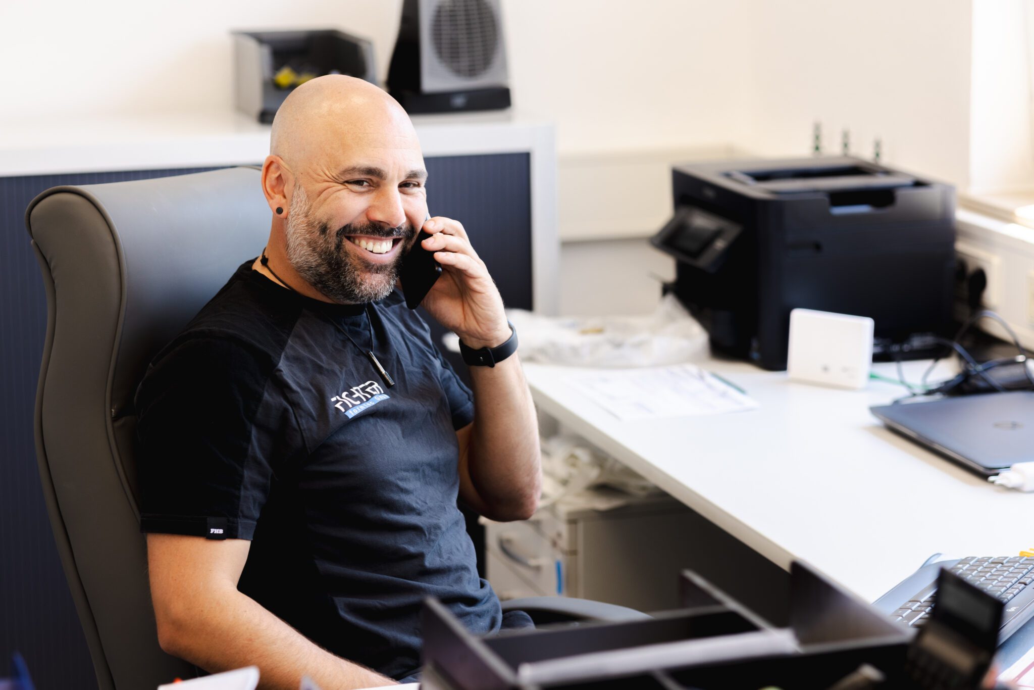 Mitarbeiter im Büro führt ein Telefonat und lächelt dabei, Arbeitsplatz mit Laptop und Drucker im Hintergrund