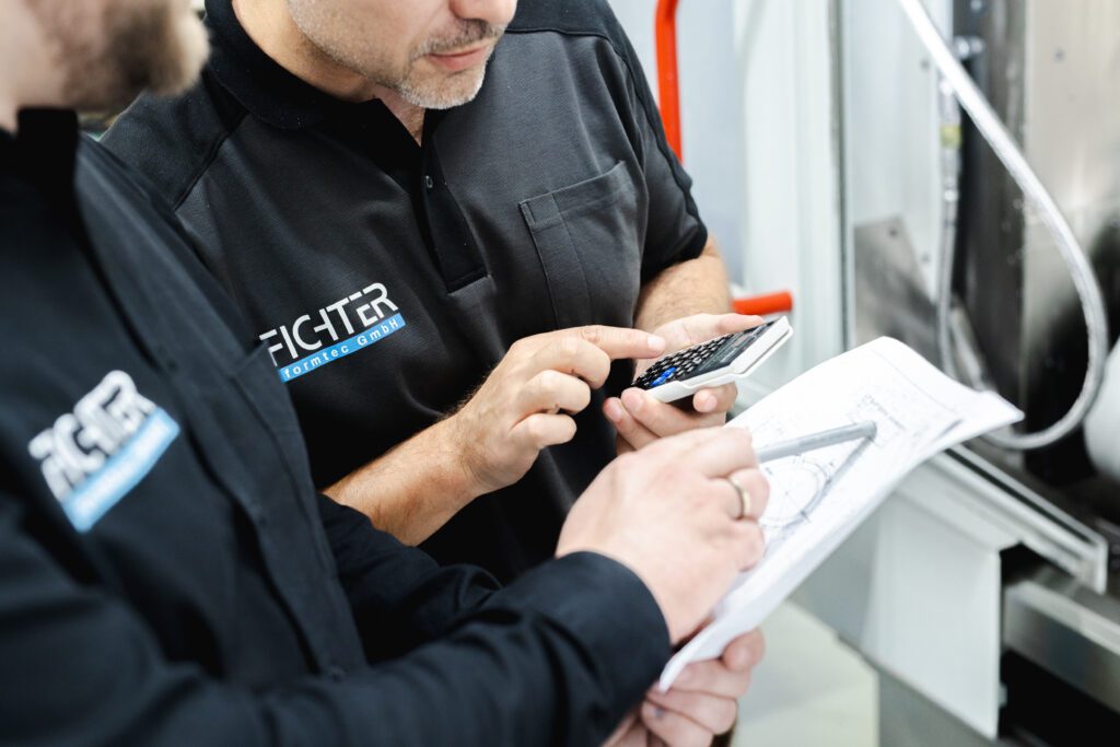 Zwei Mitarbeiter von Fichter Formtec in schwarzer Firmenkleidung prüfen technische Zeichnungen und nutzen einen Taschenrechner