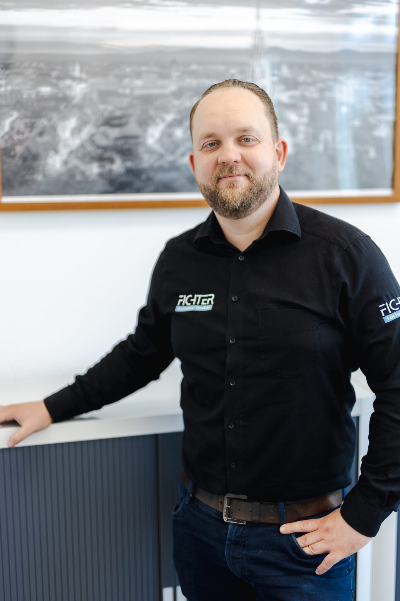 Geschäftsführer von Fichter formtec im Vertriebsbereich steht lächelnd in Firmenkleidung vor einem Schreibtisch